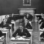 Gezeigt wird eine Schulklasse in schwarz-weiß, die an ihren Schreibpulten sitzt. Alle Schüler sind männlich und in Anzug und Krawatte gekleidet.
