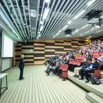 Gezeigt wird eine Vorlesung in ein einem modernen Hörsaal. Studierende sitzen Reihen. Ein Dozent steht vor ihnen. Zwei Beamer werfen Vortragsunterlagen an die Leinwände. Die Perspektive ist von der Seite.