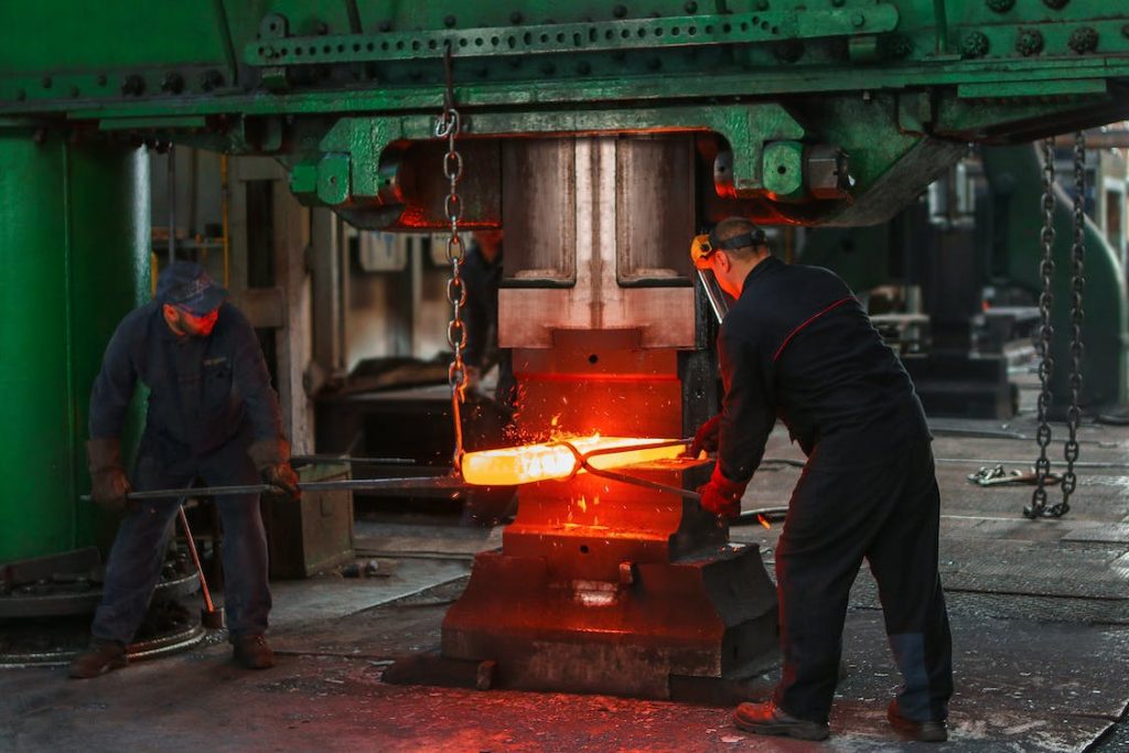 ezeigt werden zwei Männer in blauer Arbeitskleidung, die in einer modernen Werkstatt mithilfe einer großen Maschine an einem glühend heißen Werkstück arbeiten. Einer der beiden Männer trägt einen Gesichtsschutz.